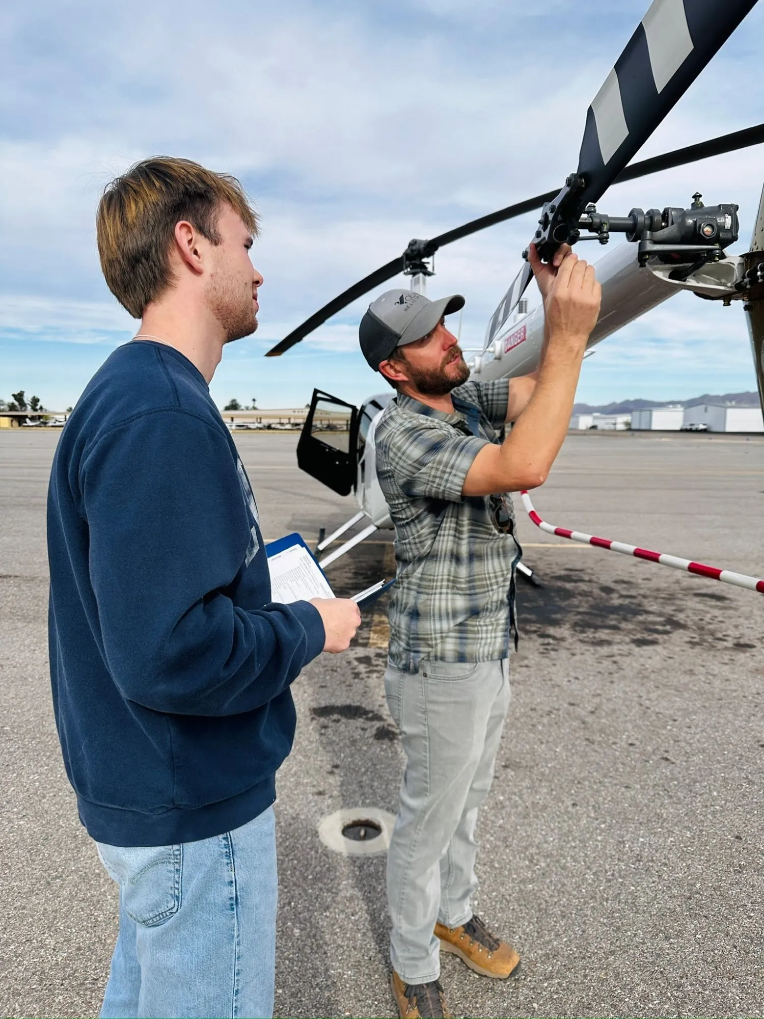 Brandon with a clipboard watching an instructor check the rotor head on a preflight