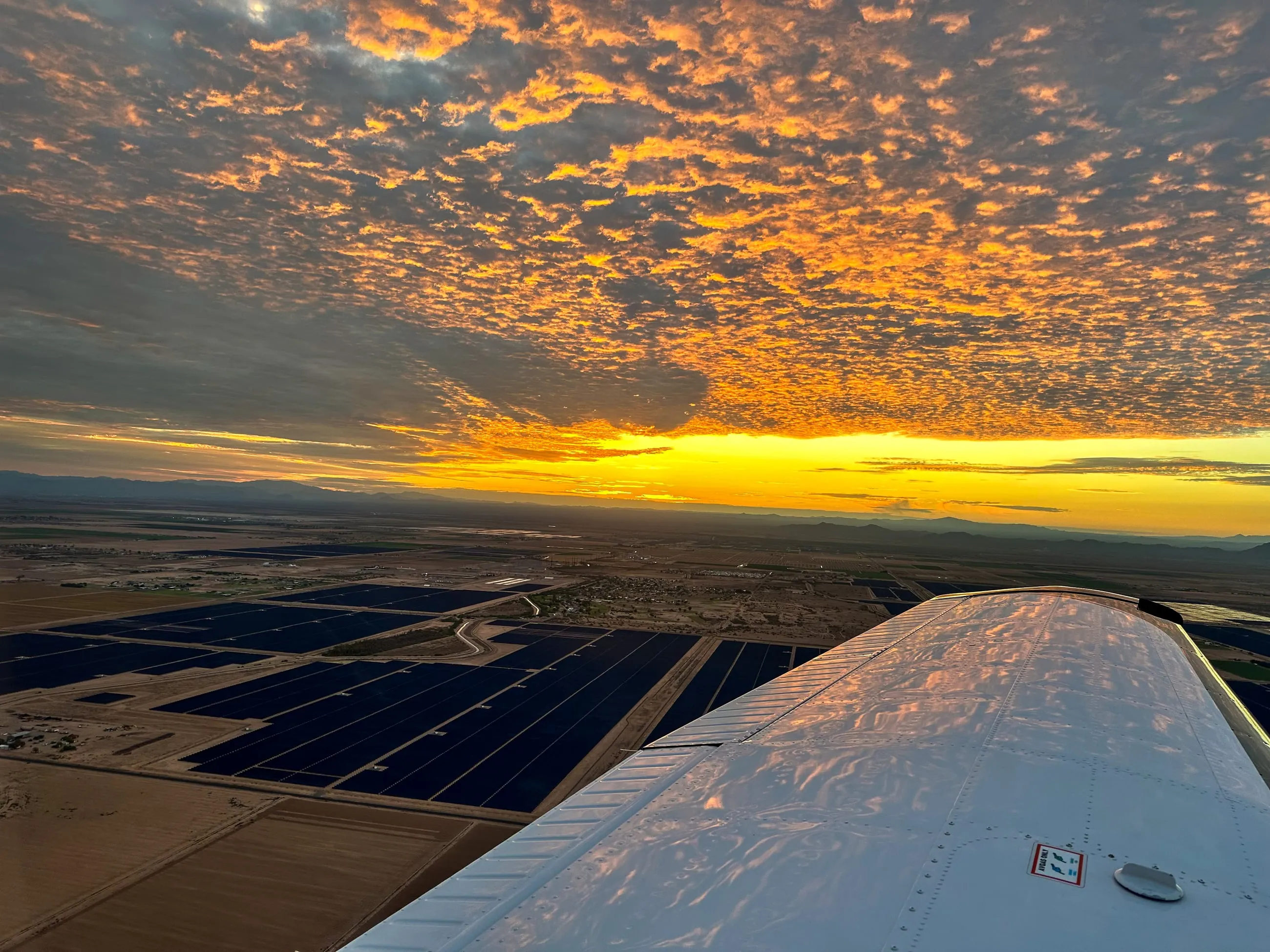 Sunset breaking under a sky of lit clouds past the wing, solar fields below